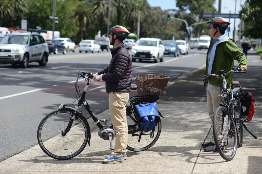 Two male adults with electric bikes waiting to cross a traffic filled road