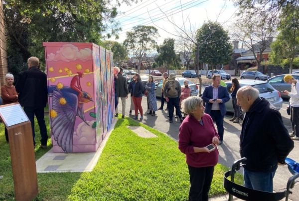 A community battery painted with a mural in pink and purple with yellow dots. People of all ages are standing around it on a sunny autumn day