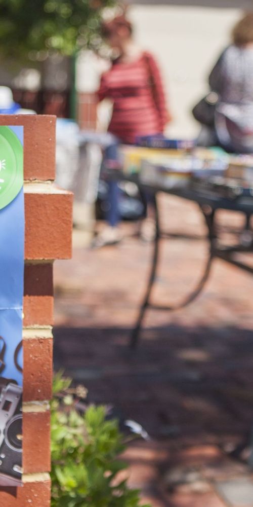 Photo of a front yard with a poster on the brick fence advertising a garage sale and people in background looking at goods on a table.