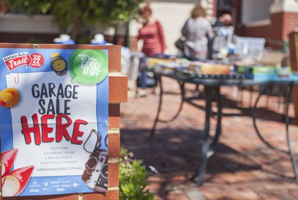 Photo of a front yard with a poster on the brick fence advertising a garage sale and people in background looking at goods on a table.