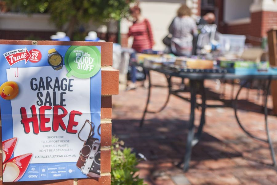Photo of a front yard with a poster on the brick fence advertising a garage sale and people in background looking at goods on a table.