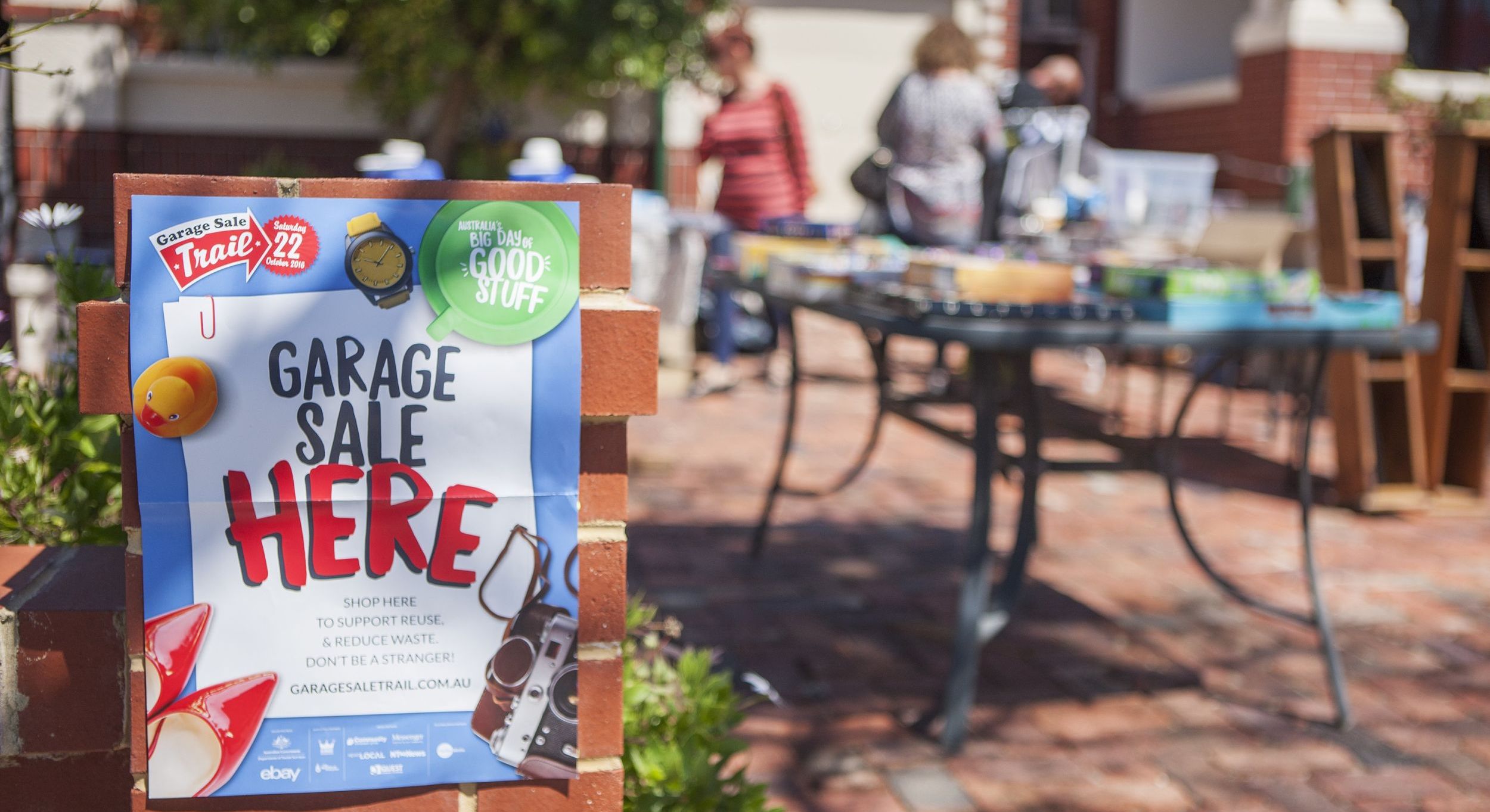 Photo of a front yard with a poster on the brick fence advertising a garage sale and people in background looking at goods on a table.