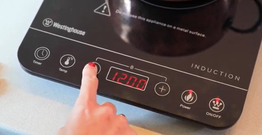 A hand pushing a button on a portable induction cooktop sitting on a bench.