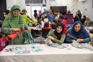 Three women with had scarves making with textiles in foreground and people in background