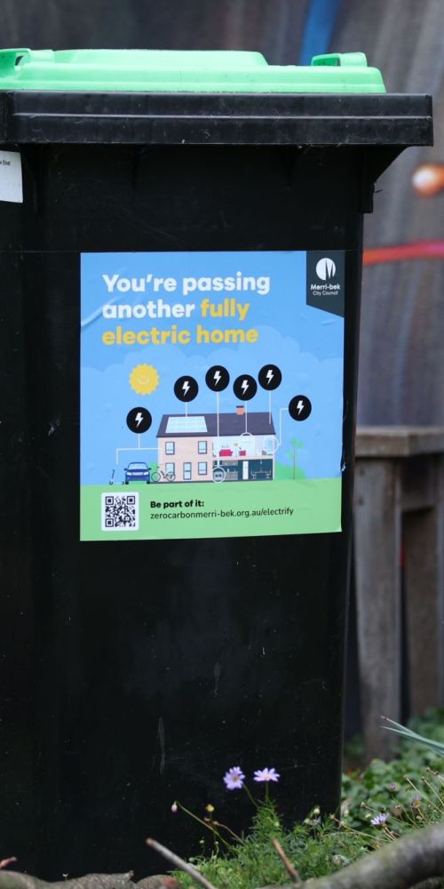 Photo of a wheelie bin with a green lid with a sticker with text "You are passing another fully electric home".