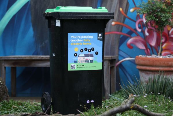Photo of a wheelie bin with a green lid with a sticker with text "You are passing another fully electric home".