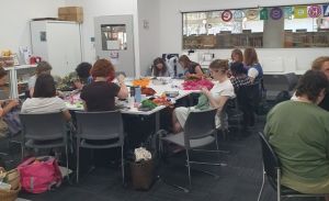 Group of people in a classroom sitting around a large stable mending their own clothes.