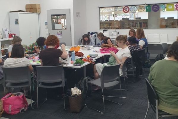 Group of people in a classroom sitting around a large stable mending their own clothes.