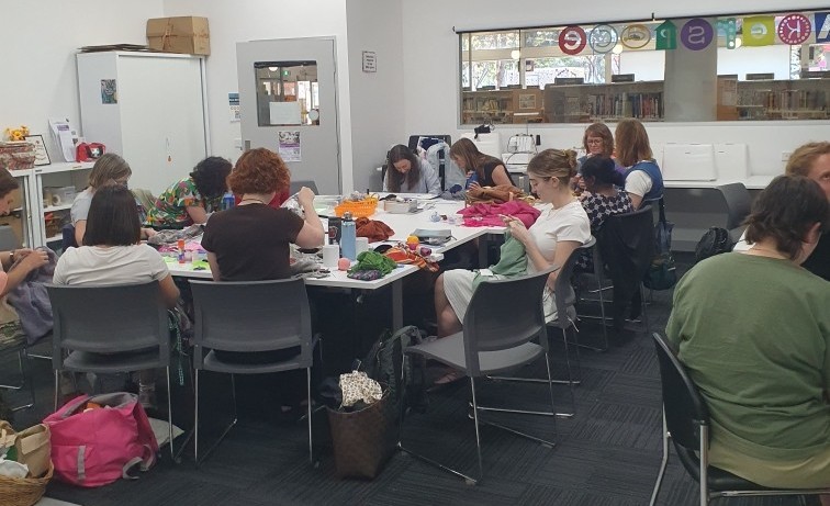 Group of people in a classroom sitting around a large stable mending their own clothes.