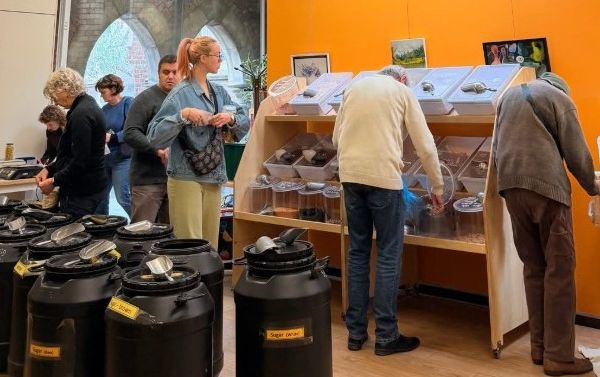 Photo with drums full of food in foreground and people walking into a room and scooping food into containers